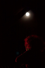 picture of the back of a person under a bridge in the dark of night illuminated by a red glow with the moon visible in the sky